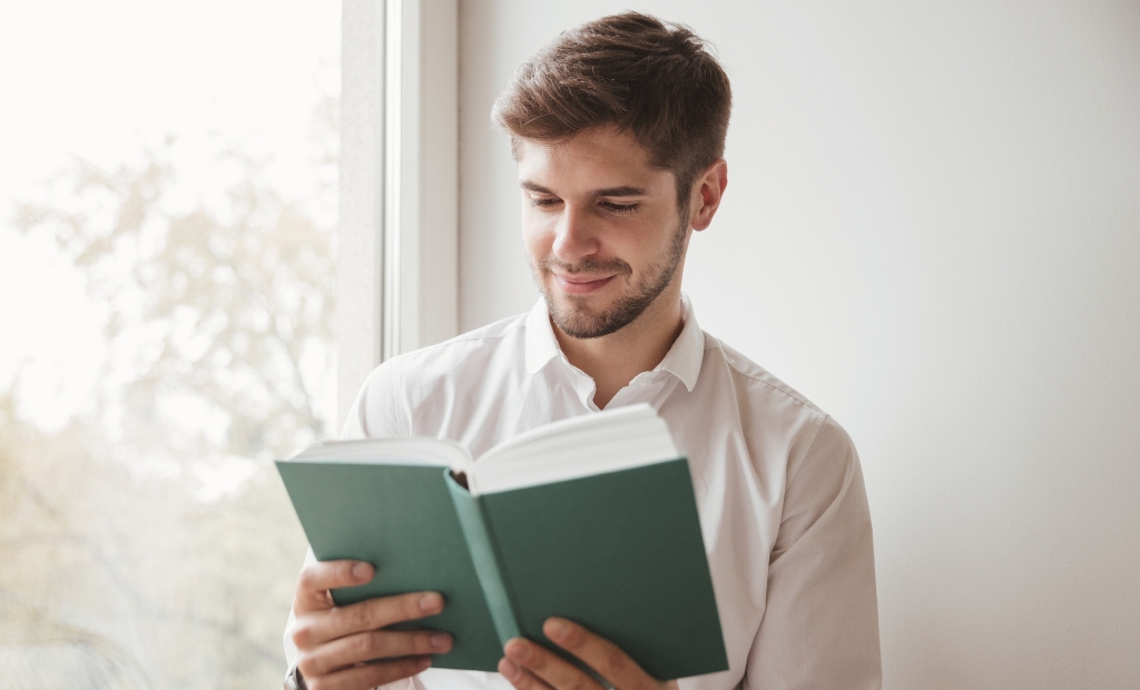 Man in a white shirt reads a green hardcover book by a sunlit window.