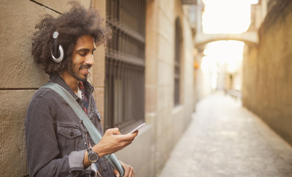 Young man with over-ear headphones leaning against a brick wall, looking at his smartphone in a sunlit alley.